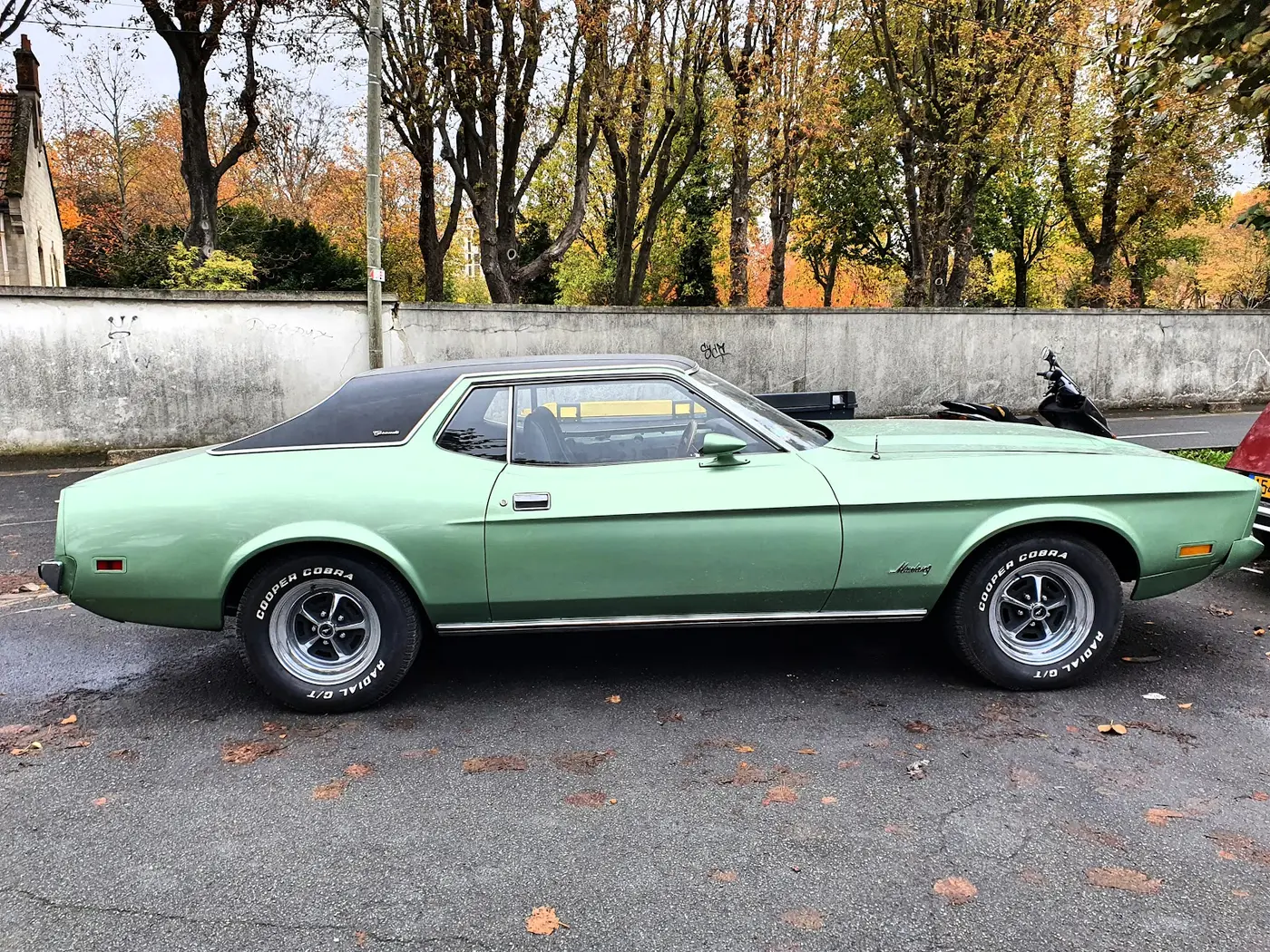 Une Ford Mustang vert amande des années 1970, garée devant un mur, vue de profil.